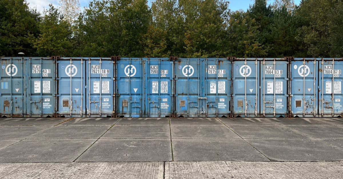Row of blue shipping containers parked along a concrete lot with green trees behind it.