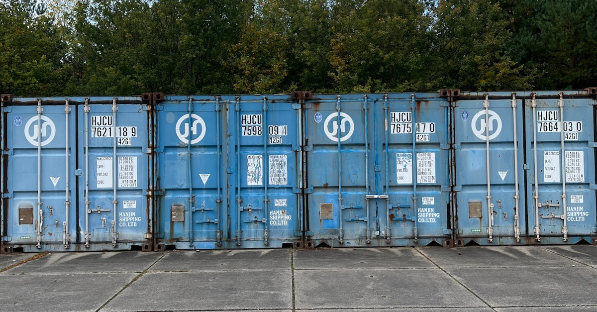 Row of blue shipping containers parked on a concrete lot with a green tree line in the background.