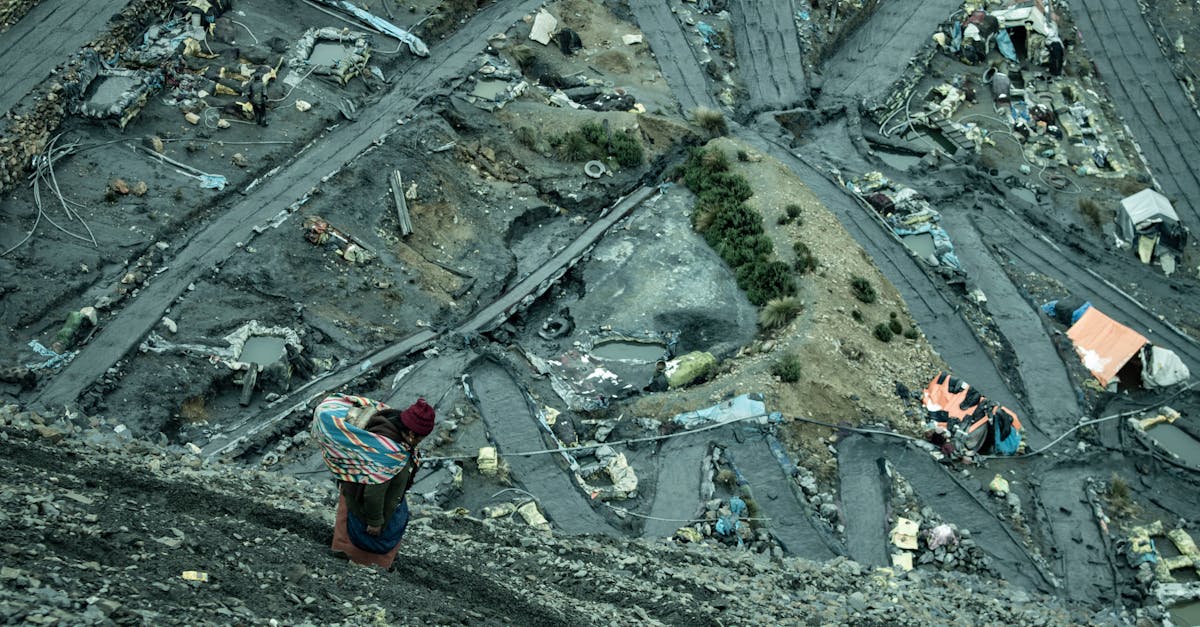 Person wearing a red hat carries a colorful bundle up a steep, debris-strewn slope overlooking a ruined site with rubble and makeshift shelters nearby.