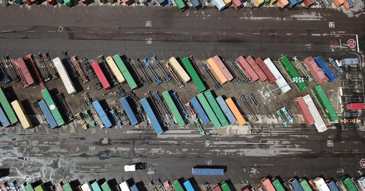 Aerial view of a shipping yard with colorful containers lined up along two long rows on a dark tarmac road in between tank-like lanes.