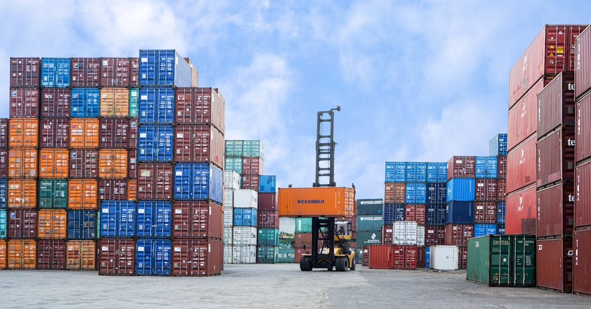 Forklift lifts an orange shipping container amid tall stacks of colorful cargo containers in a port yard.