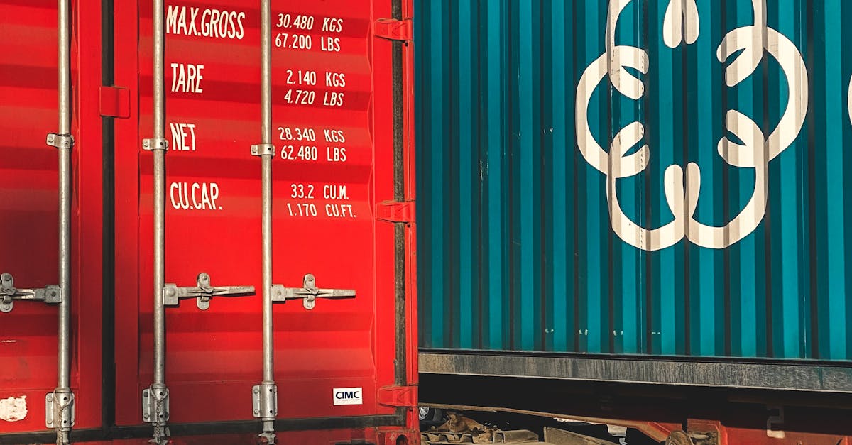 Two cargo containers side by side: a red container with white measurement text and vertical locking bars, next to a teal blue container with a large white circular logo on its side.