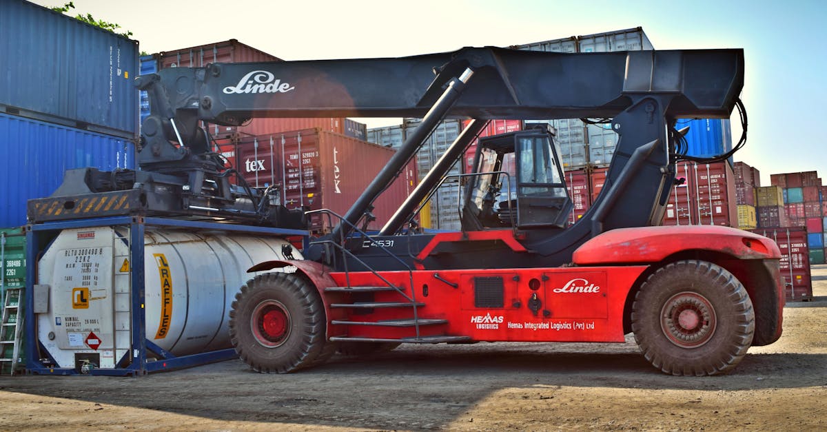 Red Linde container handler in a shipping yard beside stacked cargo containers on a dusty ground