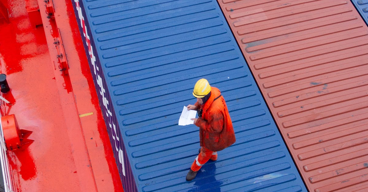 Worker in orange safety gear and a yellow hard hat walks on a blue shipping container deck, reviewing papers on a clipboard or document.