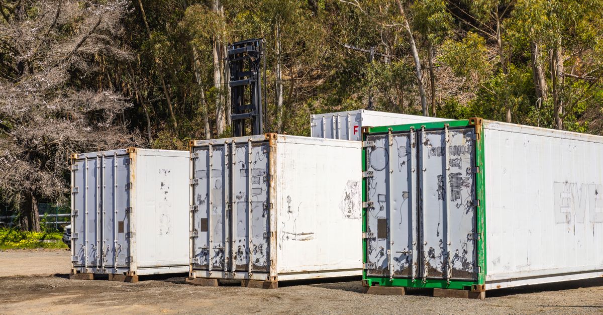Several weathered white shipping containers on dirt, with a green-framed container to the right and trees in the background.
