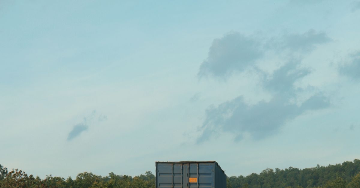 Blue sky with scattered clouds over a blue shipping container on a road, with a treeline in the distance.