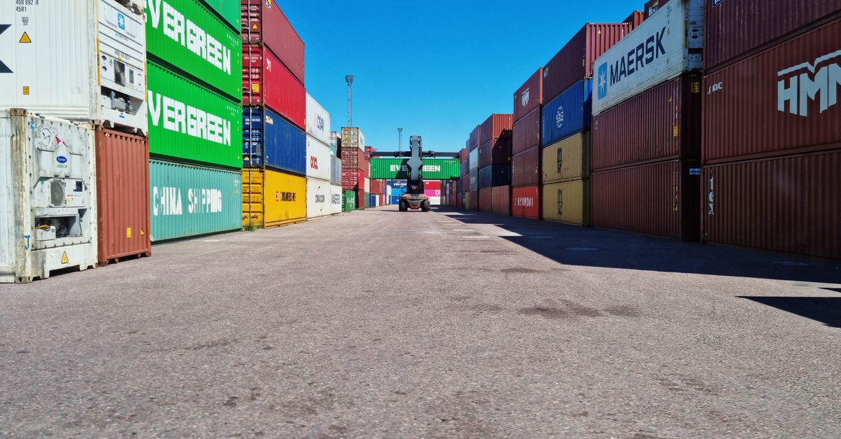 Forklift driving down a container yard with stacked colorful cargo containers on both sides under a clear blue sky.