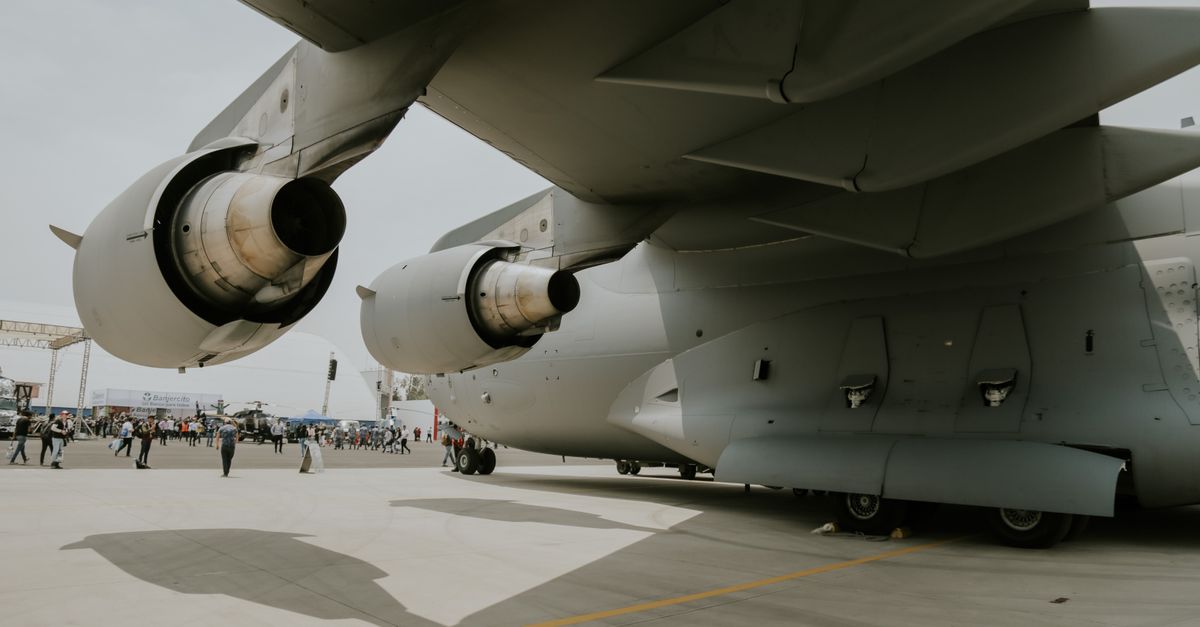 Close-up view of two large jet engines under a military transport aircraft at an airshow, with visitors in the background.