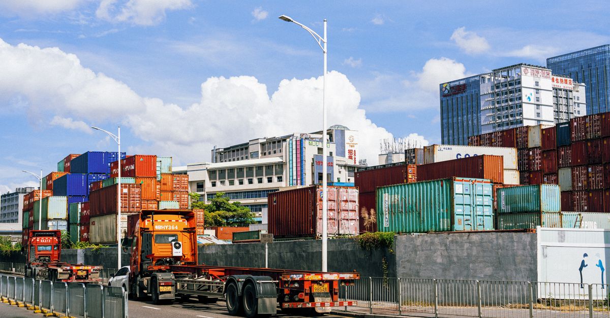 Colorful shipping containers stacked along a road with orange trucks and a flatbed trailer, urban buildings in the background under a blue sky.