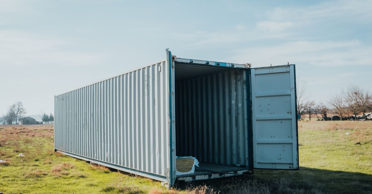 Open gray shipping container in a grassy field under a blue sky; interior dark and empty.