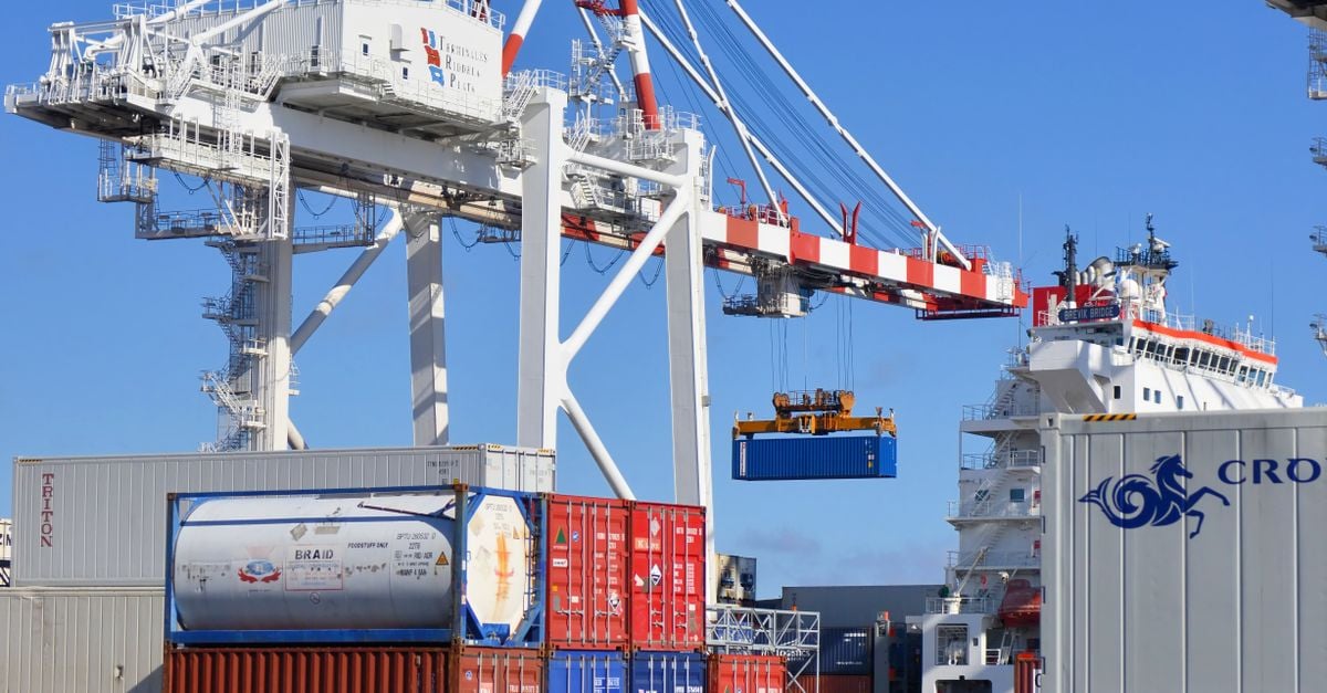 Large port container yard with towering gantry crane hoisting a blue shipping container over stacked colorful cargo containers and a docked ship nearby.