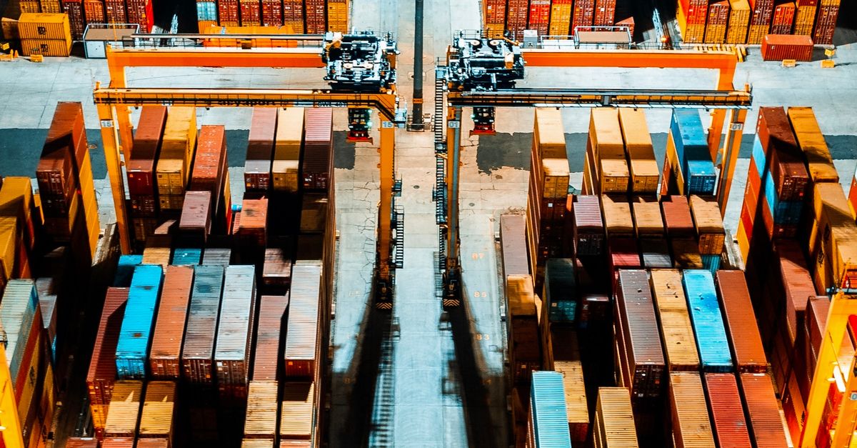 A shipping port viewed from above with rows of colorful shipping containers and two large orange gantry cranes overhead.