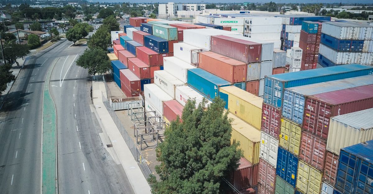 Aerial view of a large stack of colorful shipping containers beside a curved road in an industrial area.