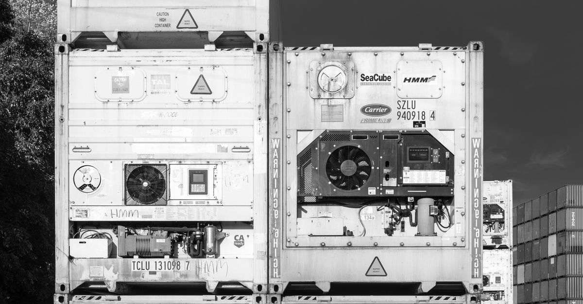 Two stacked cargo containers in a yard, showing exposed cooling equipment, vents, and warning labels in black-and-white.
