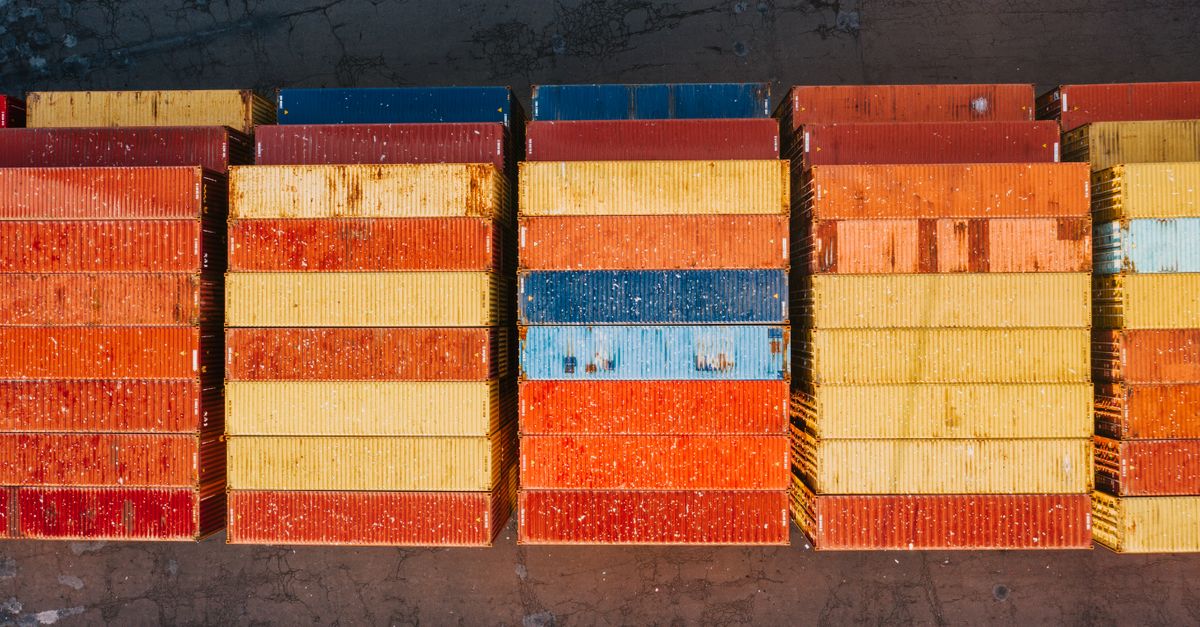 Stacks of colorful shipping containers in red, orange, yellow, and blue on a dusty surface, seen from above.