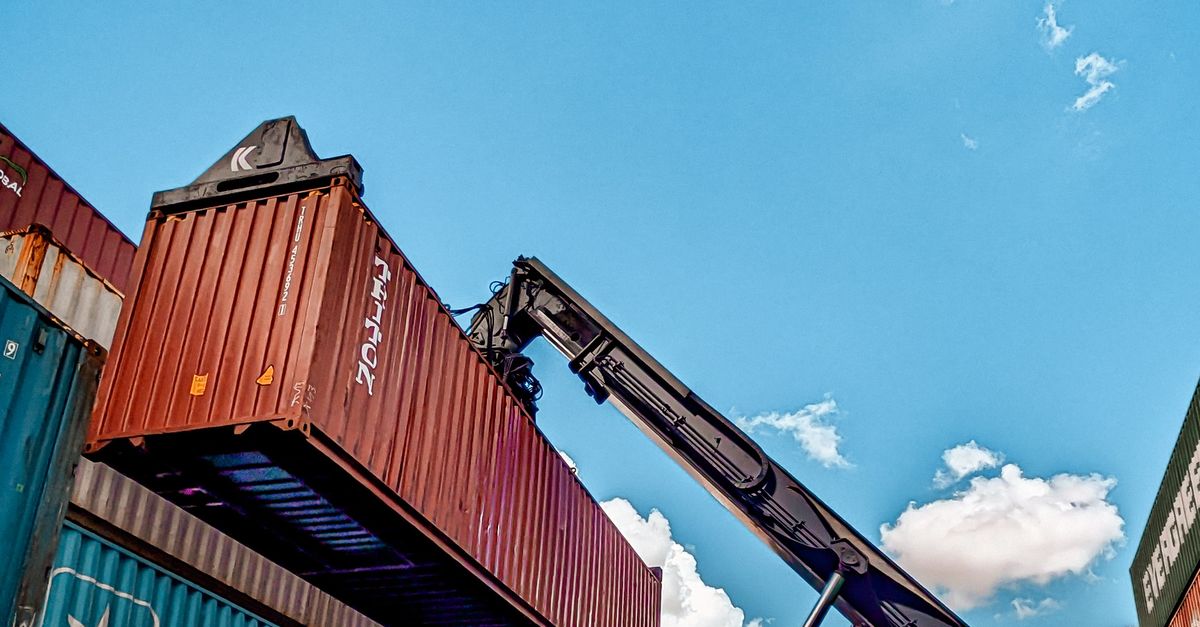 Crane lifting a red shipping container onto stacked cargo containers against a blue sky