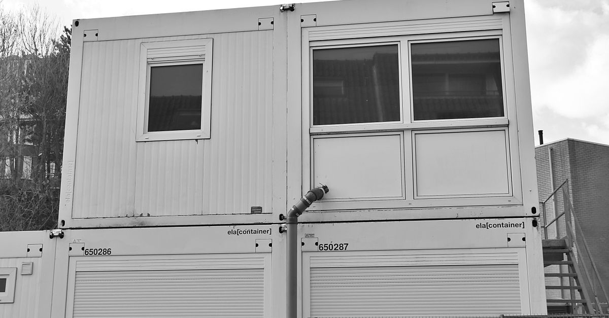 Two stacked modular shipping containers outdoors, with a small window on the upper unit and a metal pipe joining the boxes.
