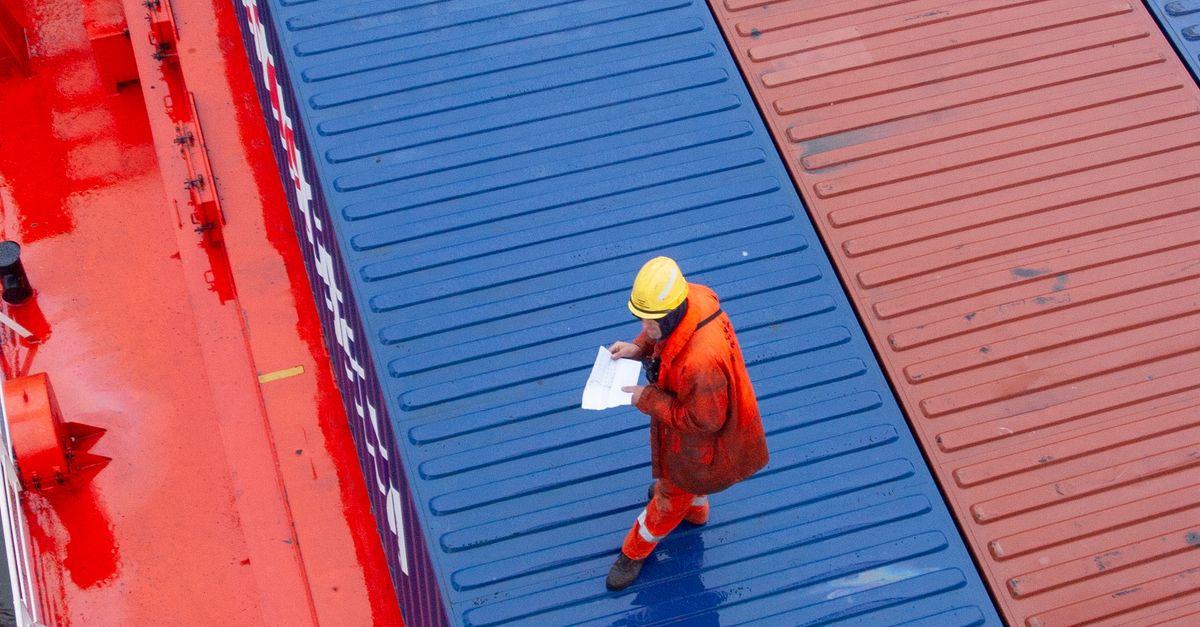 Cargo ship worker in orange suit and yellow hard hat standing on a blue shipping container, examining documents.