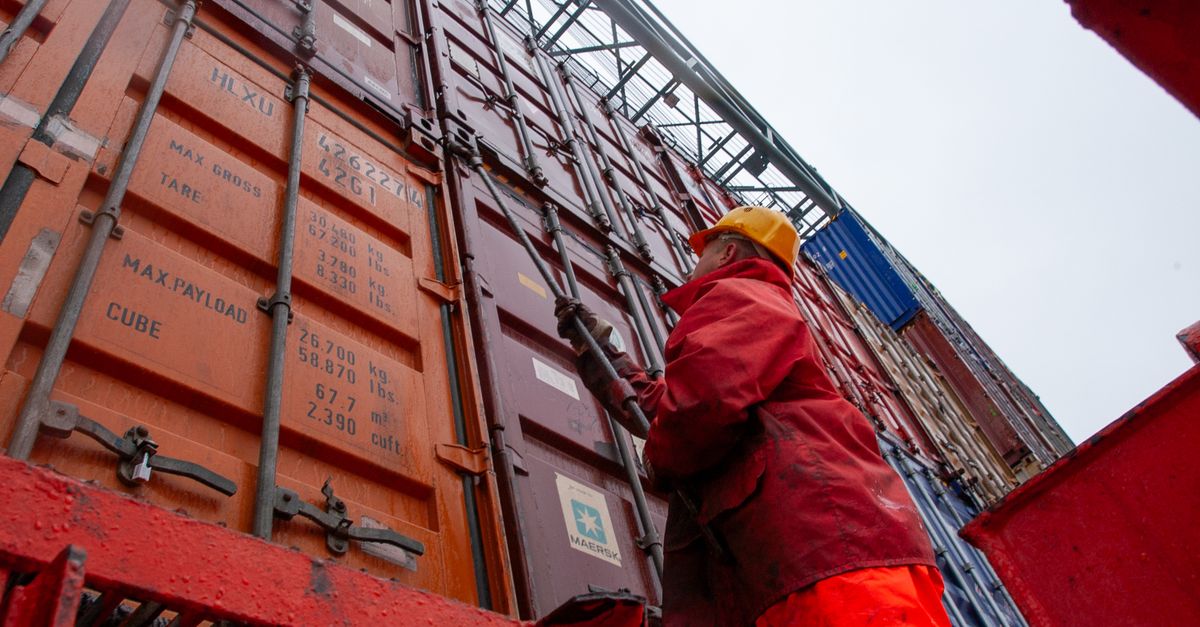 Port worker in red coveralls and yellow helmet near stacked orange and blue shipping containers.