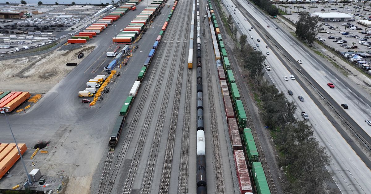 Aerial view of a freight yard with long train tracks, stacked cargo containers, and a parallel highway with cars nearby